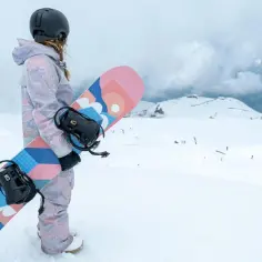 Snowboarder w kasku i kolorowym stroju trzyma deskę snowboardową z grafiką.