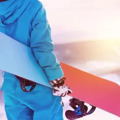 Osoba w niebieskiej kurtce i rękawiczkach trzyma snowboard z niebieskim i różowym spodem.
