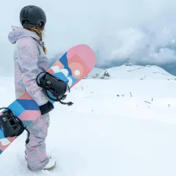Snowboarder w kasku i kolorowym stroju trzyma deskę snowboardową z grafiką.